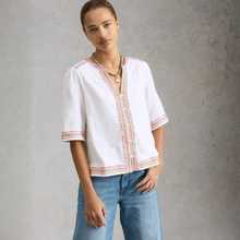Load image into Gallery viewer, Woman wearing a white blouse with red embroidery and blue jeans against a gray background
