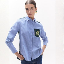 Load image into Gallery viewer, A woman modeling a light blue and white striped button-down shirt with dark green contrast panels on the shoulders and a matching pocket on the chest featuring a crest patch.
