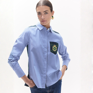 A woman modeling a light blue and white striped button-down shirt with dark green contrast panels on the shoulders and a matching pocket on the chest featuring a crest patch.