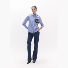 Load image into Gallery viewer, A woman modeling a light blue long-sleeve button-up shirt with a dark green and white patch on the left chest, paired with dark blue jeans and black shoes, against a plain white studio background.
