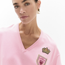 Load image into Gallery viewer, Close-up of a woman wearing a light pink V-neck sweatshirt with embroidered patches on the chest, including a crown patch and a shield patch with the text 'The Championship'.
