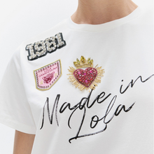 Load image into Gallery viewer, A close-up of a white short-sleeve T-shirt worn by a model, featuring decorative embellishments including a gold and pink crown heart patch, a pink shield patch, and a rhinestone '1981' patch, with 'Made in Lola' written in black script across the front.
