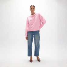 Load image into Gallery viewer, A woman modeling a light pink crewneck sweatshirt with a minimalist line-art graphic on the front, paired with blue jeans and brown pointed-toe flats, against a white studio background.
