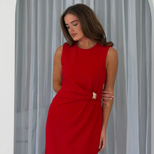 Load image into Gallery viewer, A woman wearing a sleeveless bright red dress with a gathered waist and a small white bow detail, standing in front of light gray curtains.
