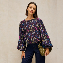 Load image into Gallery viewer, A woman modeling a long-sleeve blouse with a dark abstract floral pattern in purple, blue, pink, and beige, paired with dark blue jeans and holding a small olive green handbag.
