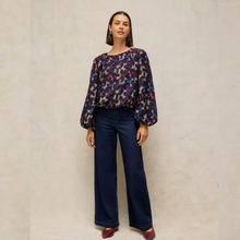Load image into Gallery viewer, A woman stands against a textured beige wall wearing a long-sleeve blouse with a purple, pink, and blue abstract floral pattern, paired with dark blue wide-leg jeans and burgundy flats.
