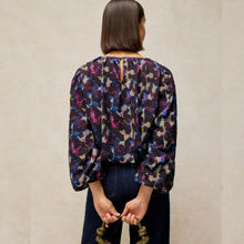 Load image into Gallery viewer, A woman viewed from behind wearing a long-sleeve blouse with a dense abstract multicolor print in purple, blue, pink, beige, and black, paired with dark blue jeans against a neutral beige textured wall.
