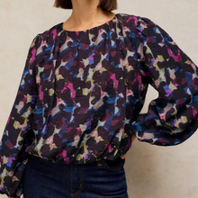 Load image into Gallery viewer, A woman wearing a long-sleeve crinkled blouse with an abstract multicolor print in black, purple, pink, blue, and beige, paired with dark blue jeans against a neutral beige background.
