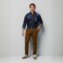 Load image into Gallery viewer, A male model stands in a studio wearing a dark blue denim button-down shirt, brown trousers, a black belt, and sneakers against a light gray background.
