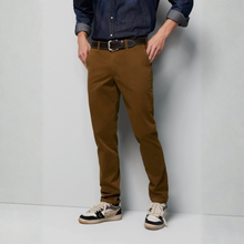 Load image into Gallery viewer, A person wearing slim-fit brown chino pants, a dark blue denim shirt, a black belt, and beige sneakers, standing against a light gray studio wall.
