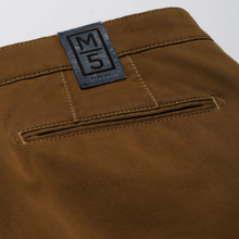 Load image into Gallery viewer, Close-up of the back waistband of brown trousers, showing a blue woven label with a black 'M5' logo and a welt pocket with contrast stitching.
