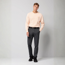 Load image into Gallery viewer, A male model stands in a studio wearing a light peach crewneck sweatshirt, dark gray trousers, a black belt, and black sneakers against a light gray background.
