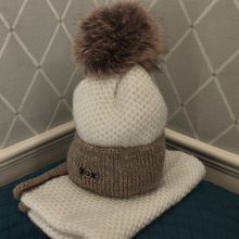 Load image into Gallery viewer, A knit winter hat with a fluffy pom-pom, shown folded on a quilted surface against a paneled wall. The hat has a cream textured upper section, a taupe-brown ribbed lower section with a small decorative emblem, and a matching brown pom-pom.
