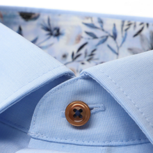Load image into Gallery viewer, Close-up of a light blue shirt with a brown button and floral pattern in the background
