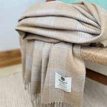 Load image into Gallery viewer, A folded woven throw blanket with a beige and off-white check pattern and fringe, draped over a wooden chair. A white brand label reading 'MCNUTT Co.
