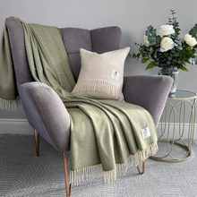 Load image into Gallery viewer, An olive green knit throw blanket with cream fringe is draped over a gray armchair, styled alongside a beige fringed pillow and a side table with white roses.
