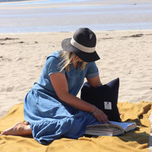 Load image into Gallery viewer, Woman in blue dress and black hat reading a book on a yellow blanket at the beach.
