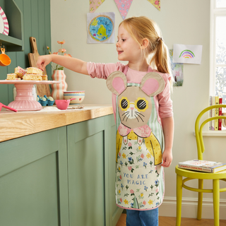 Child in a kitchen wearing a mouse-themed apron, reaching for a cake stand.