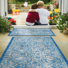 Load image into Gallery viewer, Two children sitting on a blue patterned rug in a garden setting with flowers and plants around.
