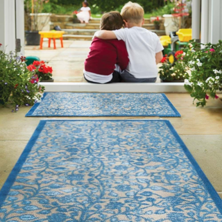 Two children sitting on a blue patterned rug in a garden setting with flowers and plants around.