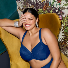Load image into Gallery viewer, A woman wearing a dark blue lace-trimmed bra and matching underwear, sitting on a mustard yellow armchair against a patterned wall.
