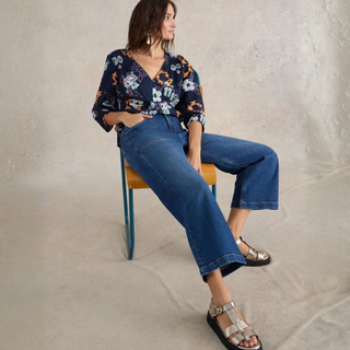 Woman sitting on a chair wearing a floral blouse and blue jeans against a neutral background