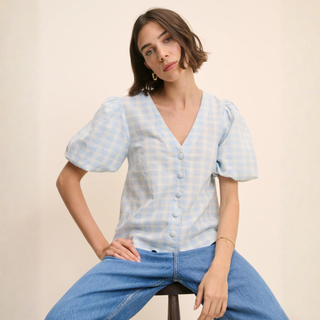 Woman wearing a light blue checkered blouse and blue jeans sitting on a stool against a beige background