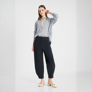 A woman modeling a blue and white vertically striped button-down shirt with dark navy wide-leg trousers and beige flat shoes, standing against a light gray studio background.