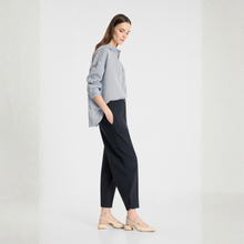 Load image into Gallery viewer, A woman stands in profile wearing a blue and white vertically striped button-down shirt, black cropped trousers, and beige flat sandals against a clean white studio background.
