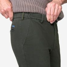 Load image into Gallery viewer, Close-up of a person wearing dark olive green chinos, showing the side pocket with a small blue brand tag, belt loops, and hands adjusting the waistband.
