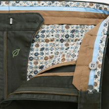 Load image into Gallery viewer, Close-up of the interior waistband and front opening of a pair of trousers, showing a dark olive green outer fabric, tan waistband, light blue inner trim, and a cream patterned lining with blue and brown motifs.
