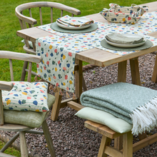 Load image into Gallery viewer, Outdoor setting with a wooden table and chairs covered with a floral tablecloth and green blanket.
