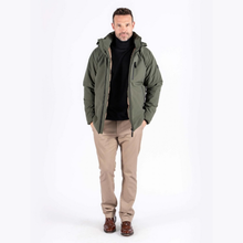 Load image into Gallery viewer, A man wearing an olive green hooded puffer jacket over a black turtleneck sweater, paired with beige trousers and brown leather shoes, standing against a plain white studio background.
