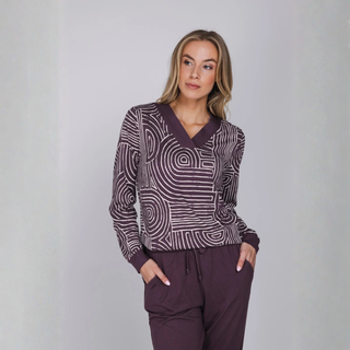 A woman modeling a long-sleeve knit top and matching pants set in a studio setting. The top features a dark plum purple and white spiral pattern, paired with solid dark plum purple trim and matching pants.