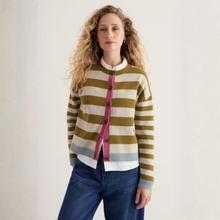 Load image into Gallery viewer, A woman with curly blonde hair wearing a striped knit cardigan with olive green, cream, and pink horizontal stripes, paired with blue jeans, standing against a plain off-white wall.
