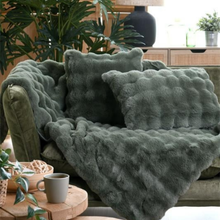 Load image into Gallery viewer, A plush, textured throw blanket and matching pillow set in a muted sage green color, styled on an armchair in a cozy living room setting with plants and a coffee mug.
