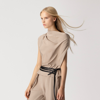 Woman wearing a beige sleeveless top and pants with a black belt on a light gray background