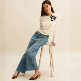 Woman wearing a white sweater with a black and yellow design and blue jeans, sitting on a wooden stool.