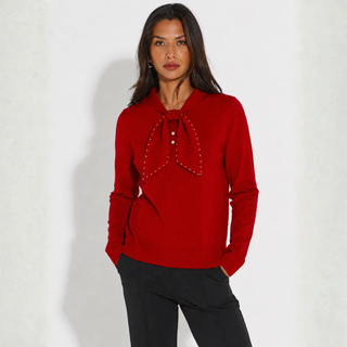 A woman modeling a bright red long-sleeve knit top with a large bow detail at the collar adorned with small silver studs, paired with black pants, against a light off-white textured wall.