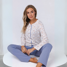 Load image into Gallery viewer, Woman sitting on a white chair wearing a patterned white shirt and light blue pants against a white background
