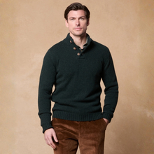 Load image into Gallery viewer, A man wearing a dark forest green knit sweater with a shawl collar and three brown buttons, paired with brown corduroy trousers, standing against a textured beige studio backdrop.
