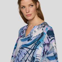 Load image into Gallery viewer, A woman wearing a loose V-neck blouse with an abstract blue, purple, and white print, photographed against a light gray studio background.

