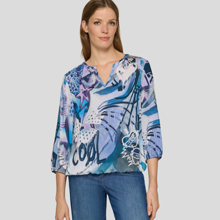 A woman modeling a blue and purple abstract print blouse with three-quarter sleeves, paired with blue jeans, against a solid light gray background.