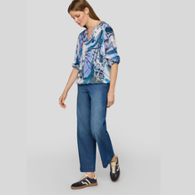 Load image into Gallery viewer, A woman modeling a blue and white abstract floral print blouse with three-quarter sleeves, paired with blue jeans and navy sneakers, against a solid light gray background.
