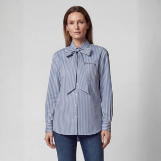 A woman modeling a blue and white vertically striped button-up shirt with a large bow tie detail at the collar and 'Prouvot' embroidered on the chest, paired with dark blue jeans against a light gray studio background.