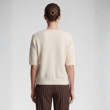 Load image into Gallery viewer, A woman viewed from the back wearing a short-sleeve cream knit sweater and brown pinstriped trousers, standing against a light gray studio background.
