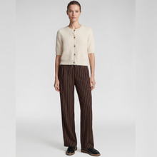 Load image into Gallery viewer, A model stands against a light gray studio background wearing a cream short-sleeve knit cardigan with buttons, brown wide-leg trousers with thin vertical stripes, and dark slip-on shoes.
