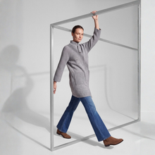 Load image into Gallery viewer, A woman wearing a gray knit turtleneck sweater and blue jeans walks through a large clear square frame with a silver metal border in a bright studio setting.
