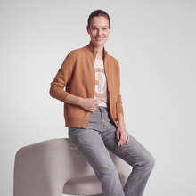 Load image into Gallery viewer, A woman sits on a light gray upholstered chair wearing a camel brown zip-front jacket over a beige graphic T-shirt and gray jeans, photographed against a plain light gray studio background.
