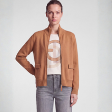 Load image into Gallery viewer, A model wearing a camel brown suede zip-up bomber jacket over a beige and brown patterned graphic t-shirt and gray jeans, set against a plain light gray studio background.
